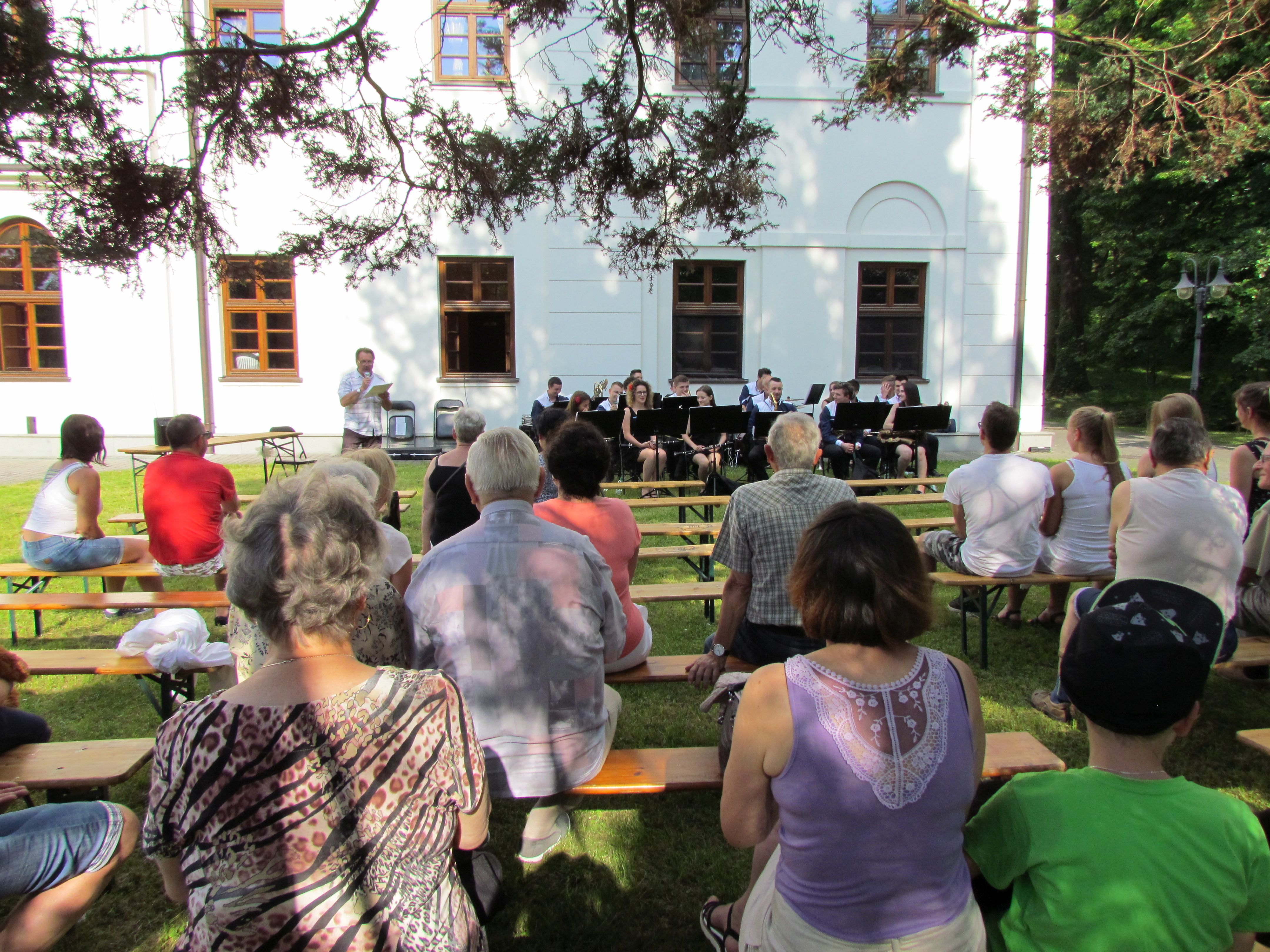 Koncert Orkiestr Dętych, widok publiczność siedzącą tyłem do obiektywu, siedzącą w kierunku  koncertującej orkiestry, w tle ściana pałacu MOW w Jaworzu